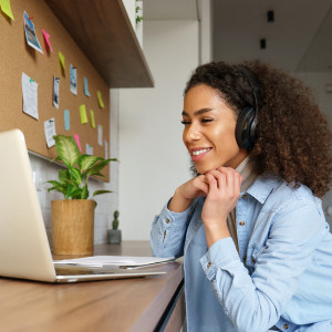 A grinning woman wearing headphones attends an online course on her laptop